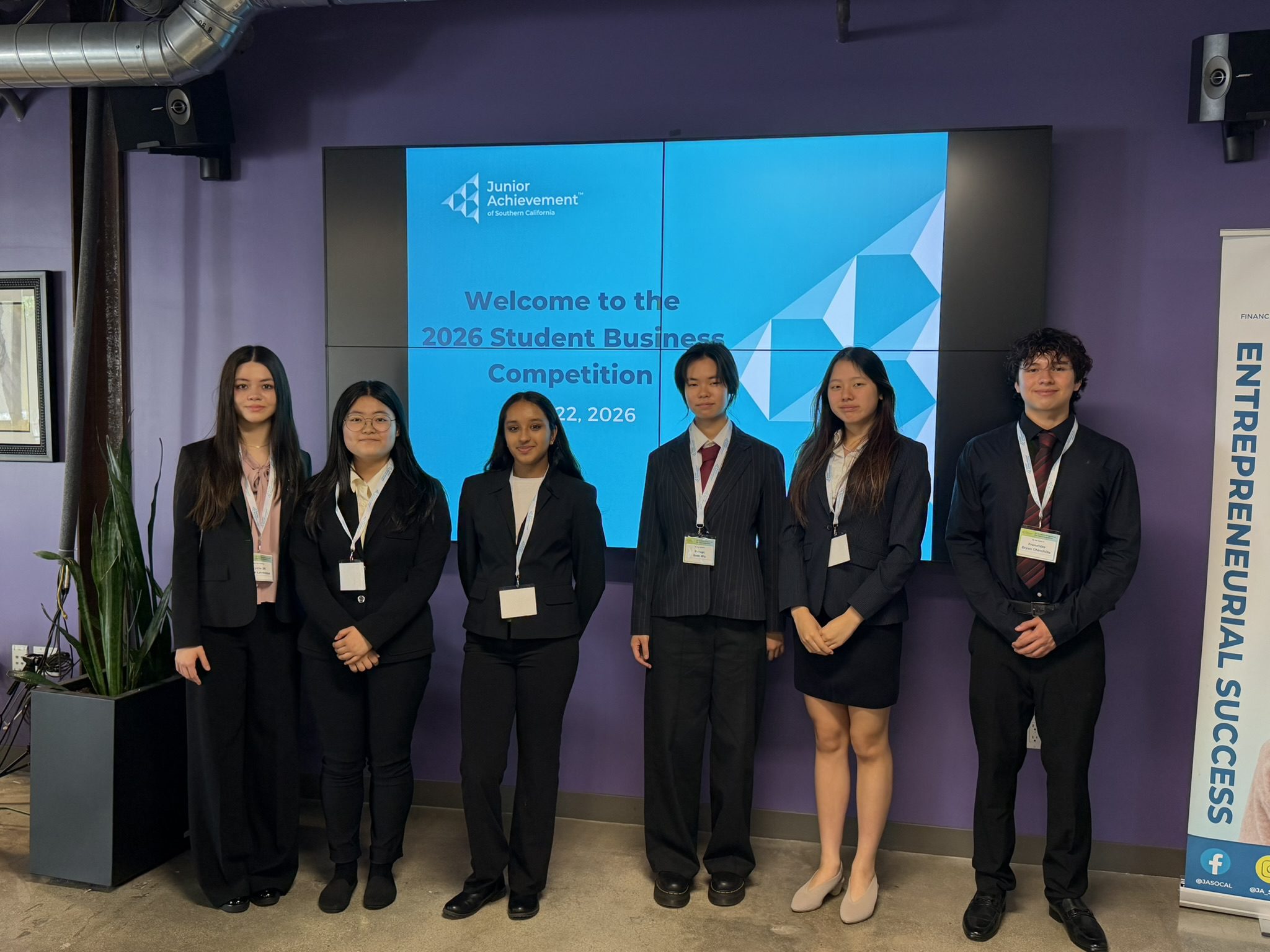 Group of six students posing in business attire in front of a blue screen that reads 'Welcome to the 2026 Student Business Competition'.