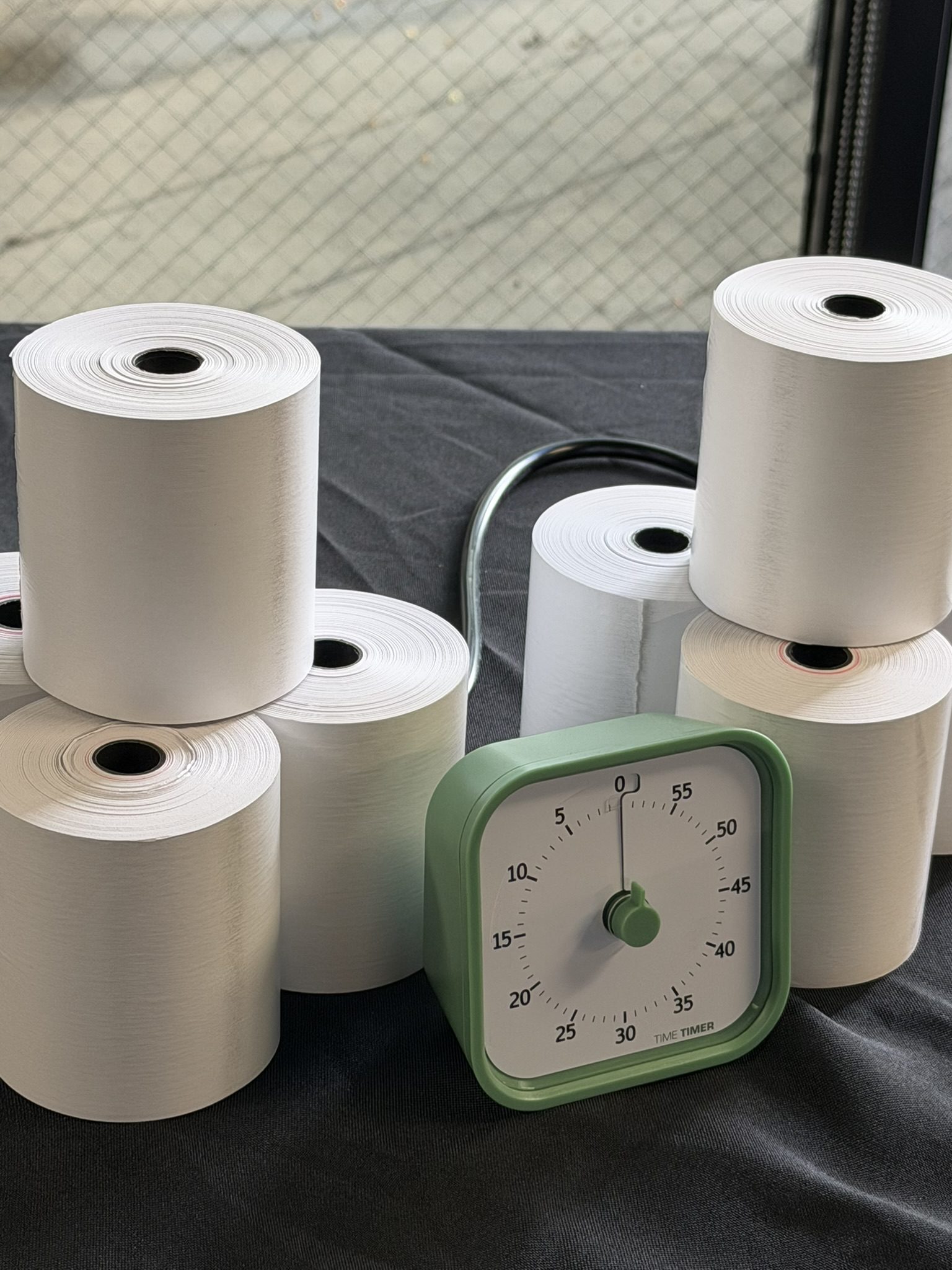 Several white paper towel rolls on a dark surface with a green kitchen timer in the foreground.