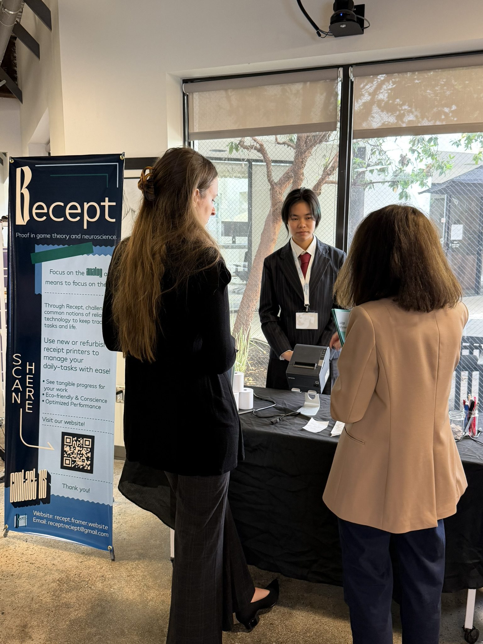 Three people stand at a booth: a presenter in a suit behind a table with a printer, talking to two visitors beside a tall banner.