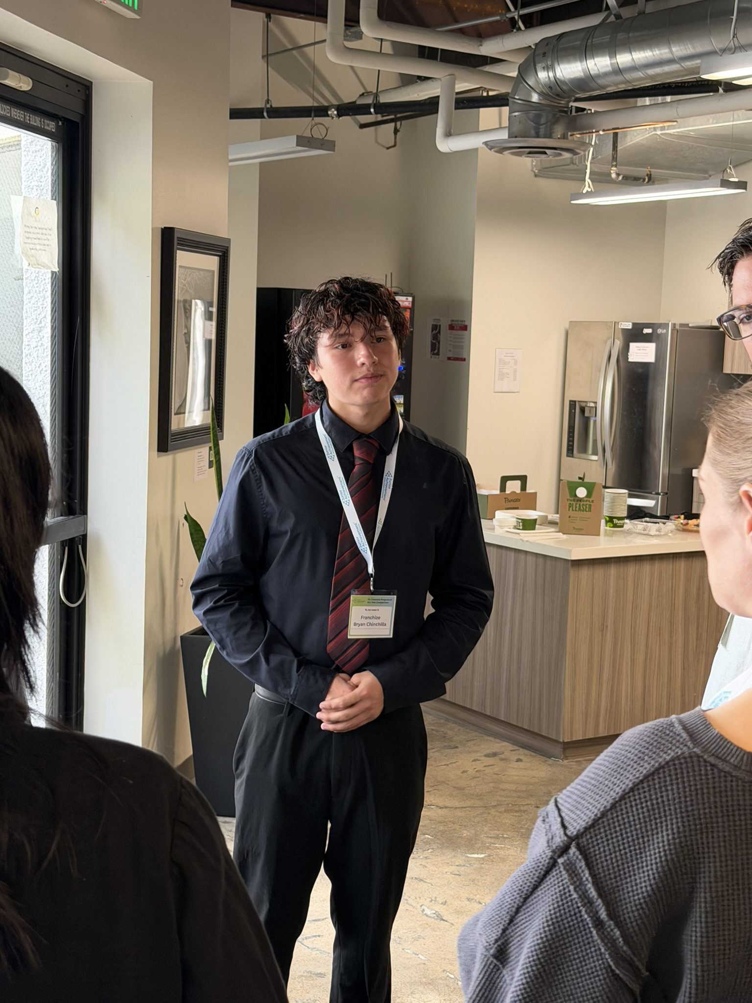 Young man in a black shirt and red striped tie with a lanyard speaks to a group in a modern office/kitchen area behind him, with a refrigerator and coffee setup visible.