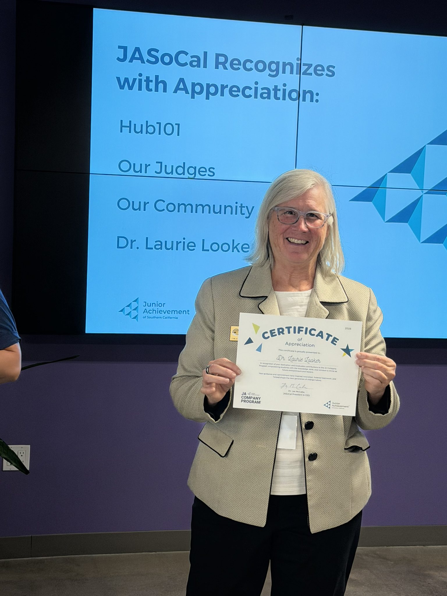 Smiling woman in a beige blazer holding a certificate of appreciation in front of a blue presentation screen.